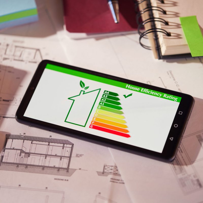 Energy efficiency screen on cellphone leaning on drawings and blueprints of a new house. Architect's desk with plans on it and a cell phone with an energy efficiency graph. Renewable energy, renovation, energy class and eco house concept.